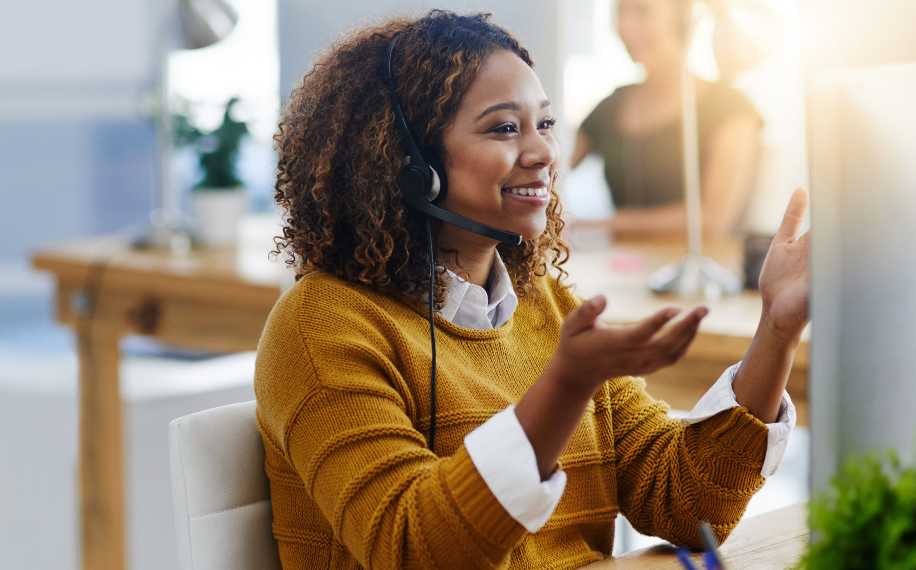 Smiling woman wearing a headset, sitting at a desk in a modern office environment, engaged in a video call