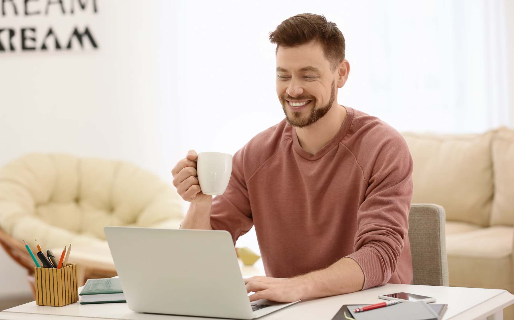 Smiling man working on a laptop at a desk while holding a coffee mug