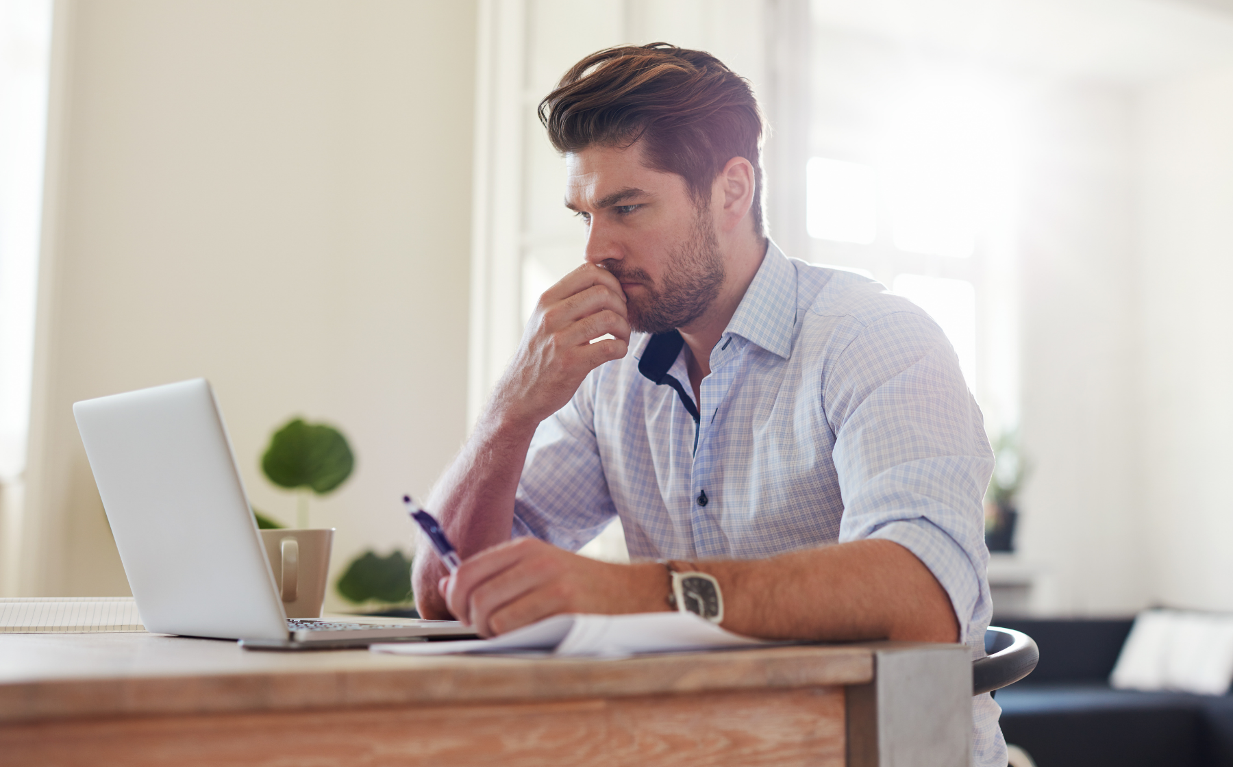 Man working at a desk, focused on a laptop while taking notes in a bright home office
