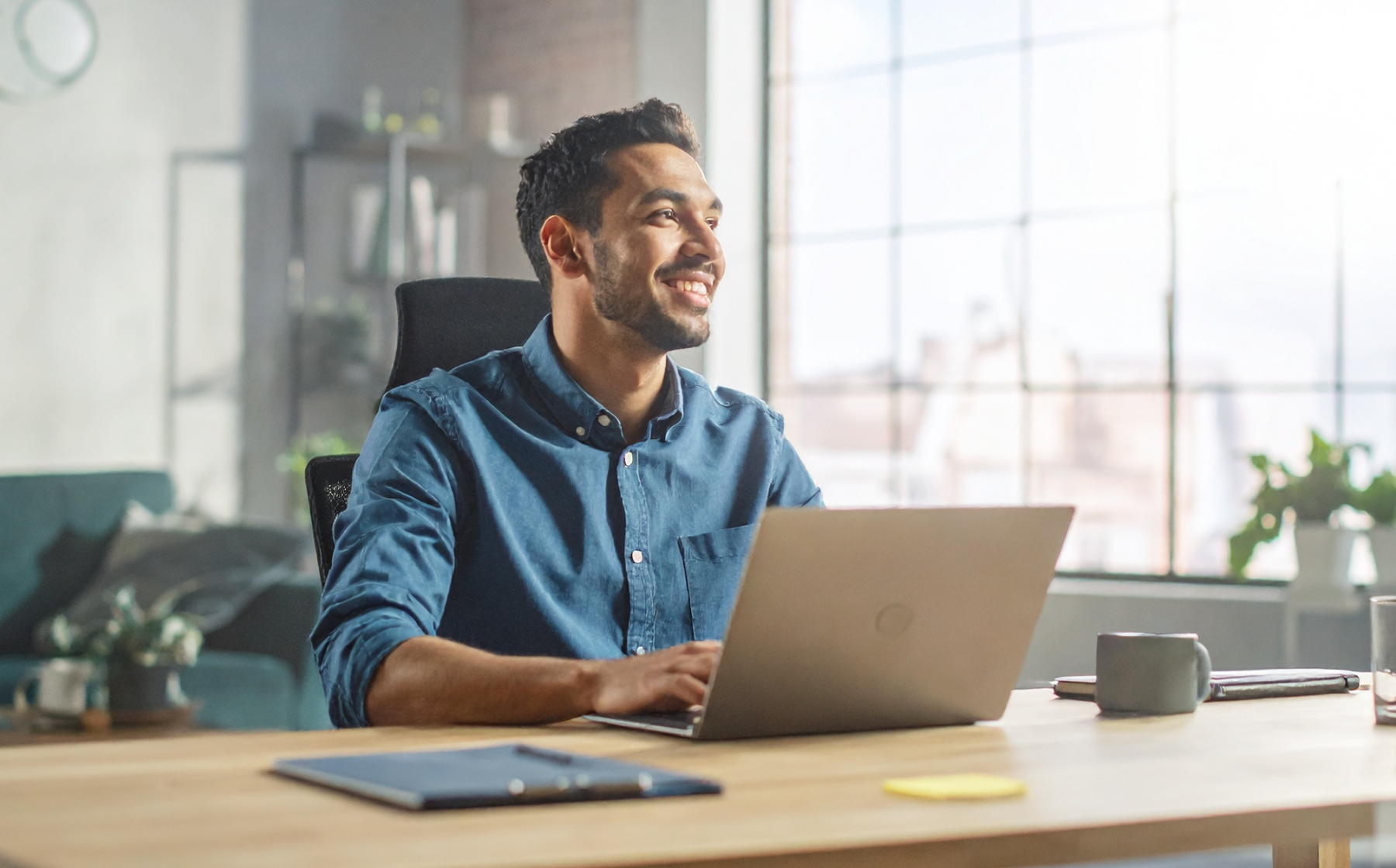 Smiling man working on a laptop at a desk in a modern, sunlit office