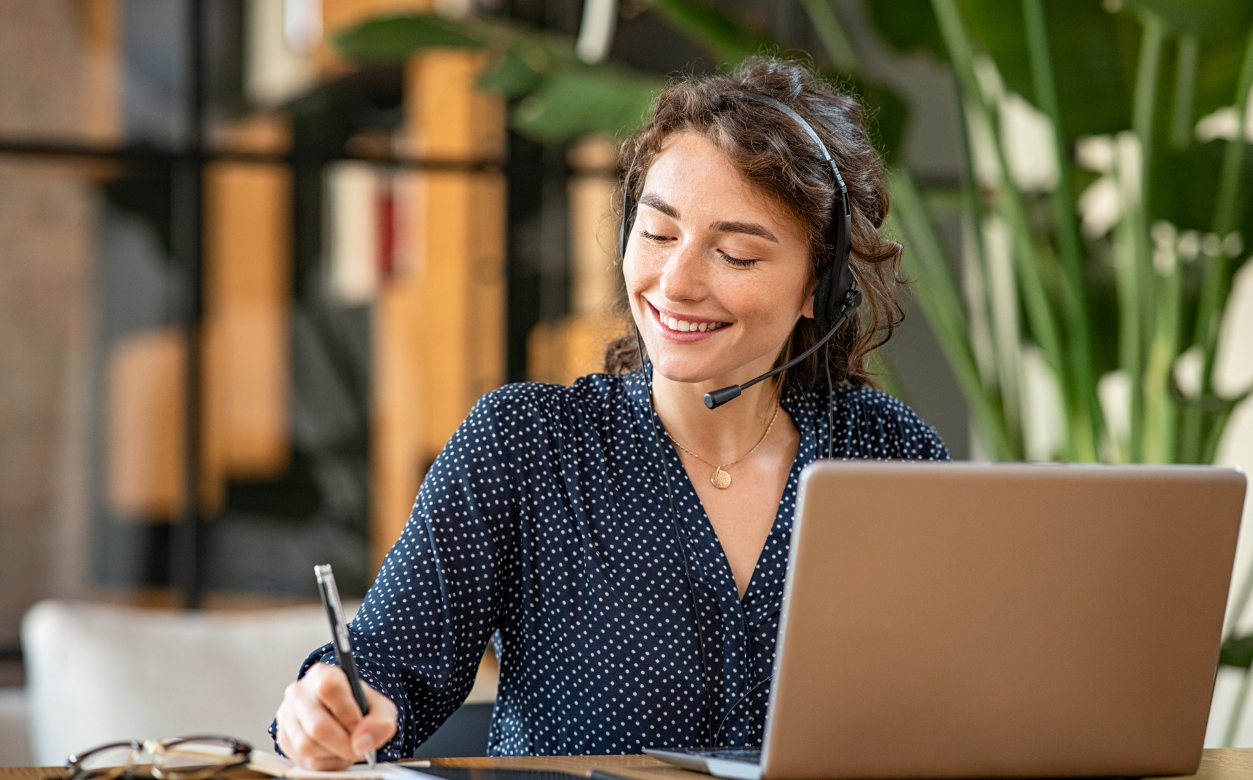 Woman with headset smiling while taking notes beside a laptop