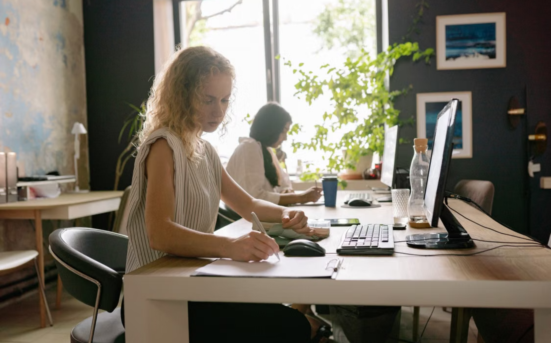 Two women work at computers in a shared office, one taking notes
